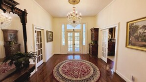 Um corredor amplo com um lustre grande e trabalhado, soalho em madeira e um tapete central.