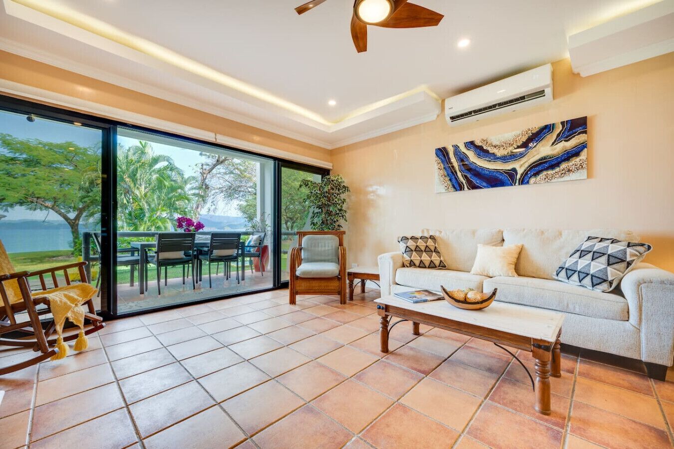 Un vaste espace de vie avec une grande porte-fenêtre coulissante donnant sur une terrasse extérieure, un canapé agrémenté de coussins à motifs, une table basse en bois et un ventilateur de plafond.
