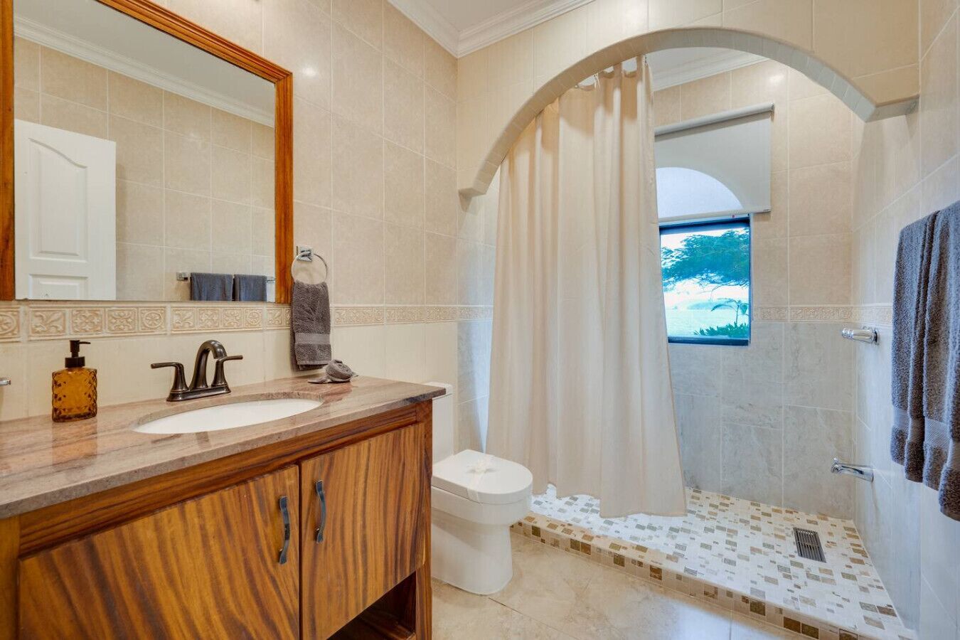 A bathroom with a wooden vanity, a sink, a mirror, and a shower area with a view of the outdoors.