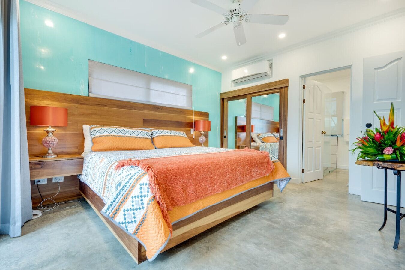Une chambre moderne avec un lit en bois, deux tables de chevet avec des lampes, un ventilateur de plafond et une vue sur un couloir.