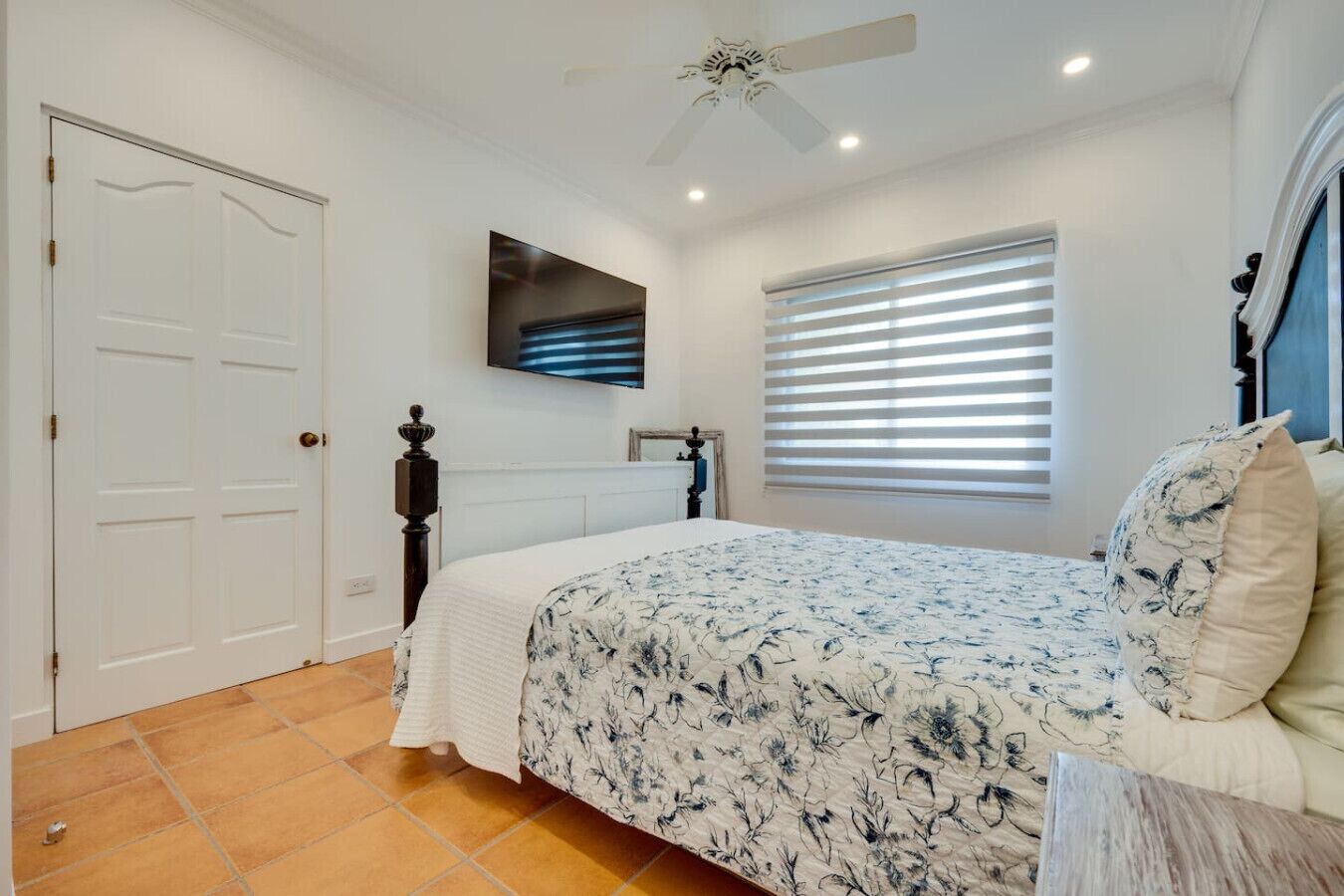A bedroom with a bed, a ceiling fan, a window with blinds, and a door.