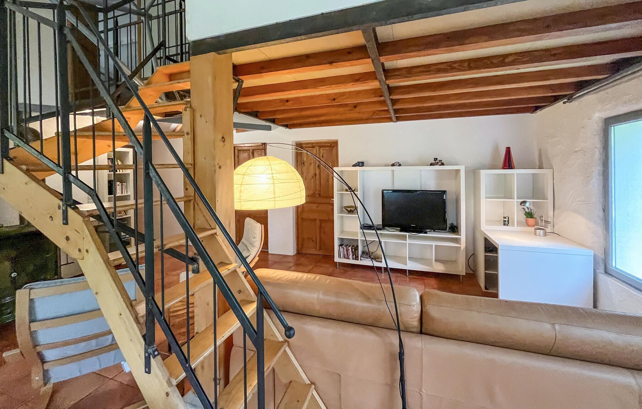 A staircase with a wooden finish and metal railings, leading to a living area with a sofa, a television, and a white shelving unit.