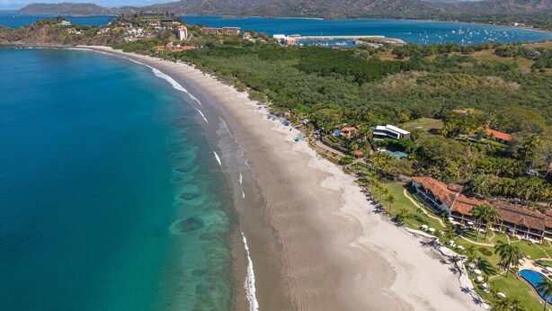 Une vue aérienne d’une zone côtière, avec une plage, un complexe hôtelier comprenant des villas, et une vue dégagée sur l’océan et les collines en arrière-plan.