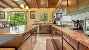 A spacious kitchen with granite countertops, wooden cabinets, and a large island. There is a dining area with a table and chairs visible through a window overlooking greenery. - Serene Mar Azul Zen Villa w/ Pool & Yoga Platform (Huacas)
