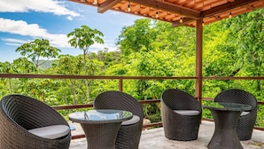 A covered outdoor seating area with wicker furniture and a glass table, overlooking a lush green landscape. - Serene Mar Azul Zen Villa w/ Pool & Yoga Platform (Huacas)