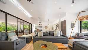 Uma sala de estar moderna com uma janela grande, um conjunto de sofás, uma mesa de centro e uma área de jantar visível pelas portas de vidro.