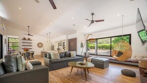 Uma sala de estar ampla com um sofá grande, uma mesa de centro e uma cadeira suspensa. Há uma área de jantar com mesa e cadeiras, e uma cozinha ao fundo.