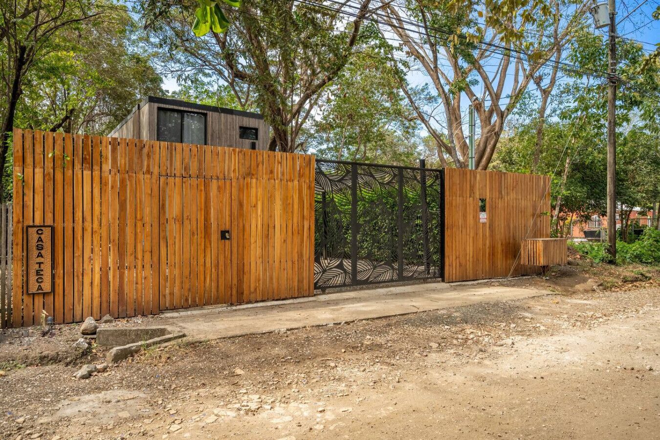 A wooden fence with a metal gate, a sign reading "CASATECA", and a dirt path leading to a building.