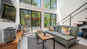 A modern living room with a grey sofa, a wooden coffee table, and a TV on the wall. There is a large window with a view of greenery and a pool outside. - Casa Teca in Surfside w/ Pool near Beach (Potrero)