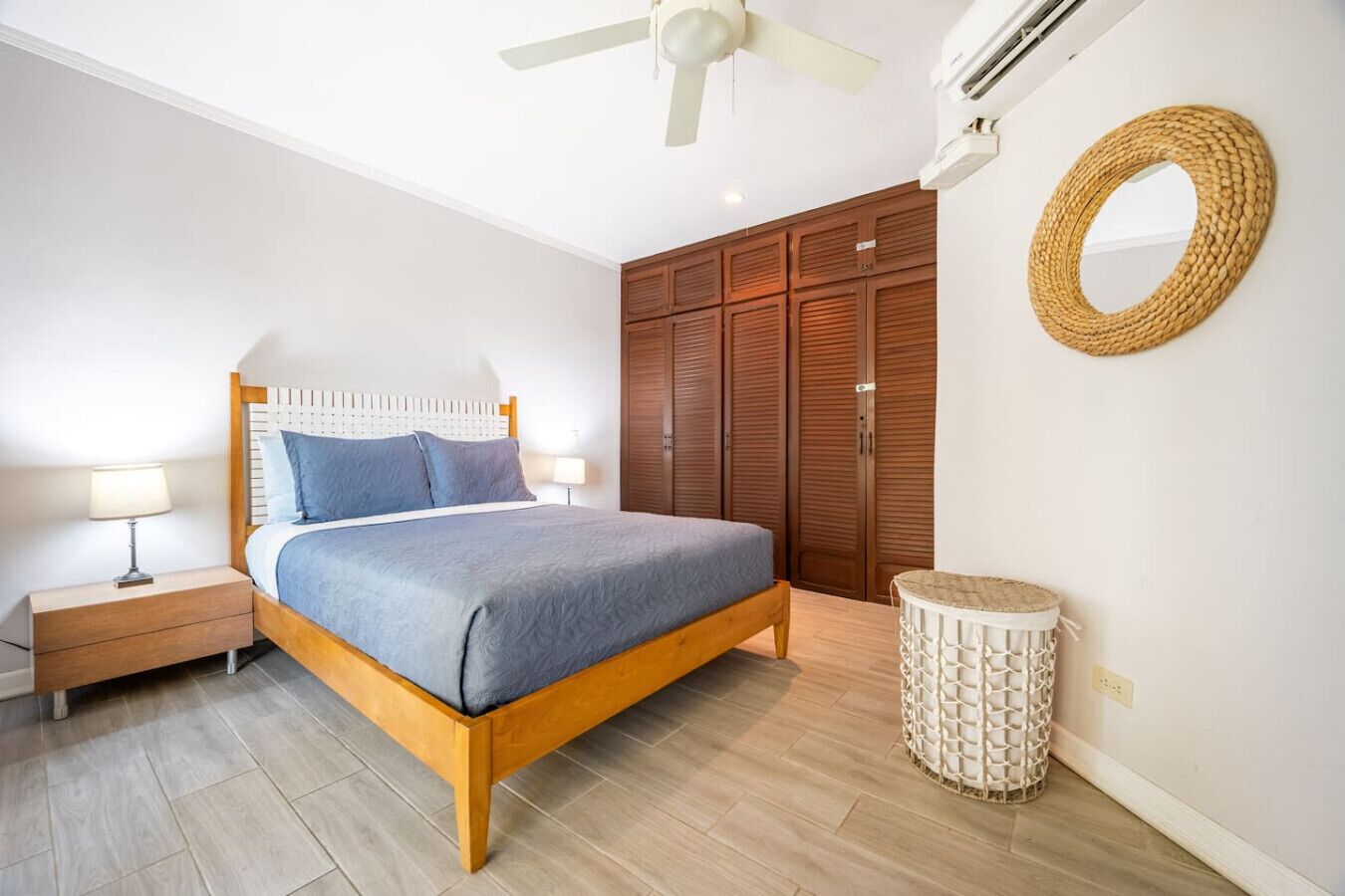 Un dormitorio con una cama de madera, dos mesitas de noche, un ventilador de techo, un armario, un espejo redondo y un taburete tejido.