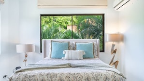 A bedroom with a large window, a bed with patterned bedding, two bedside lamps, and a decorative wall piece. - Oceanfront Condo with Pool & Stunning Views (Playa Flamingo)