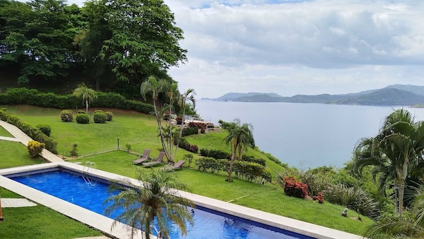 A rectangular swimming pool with a clear blue water, surrounded by well-maintained grass and palm trees. - Oceanfront Condo with Pool & Stunning Views (Playa Flamingo)