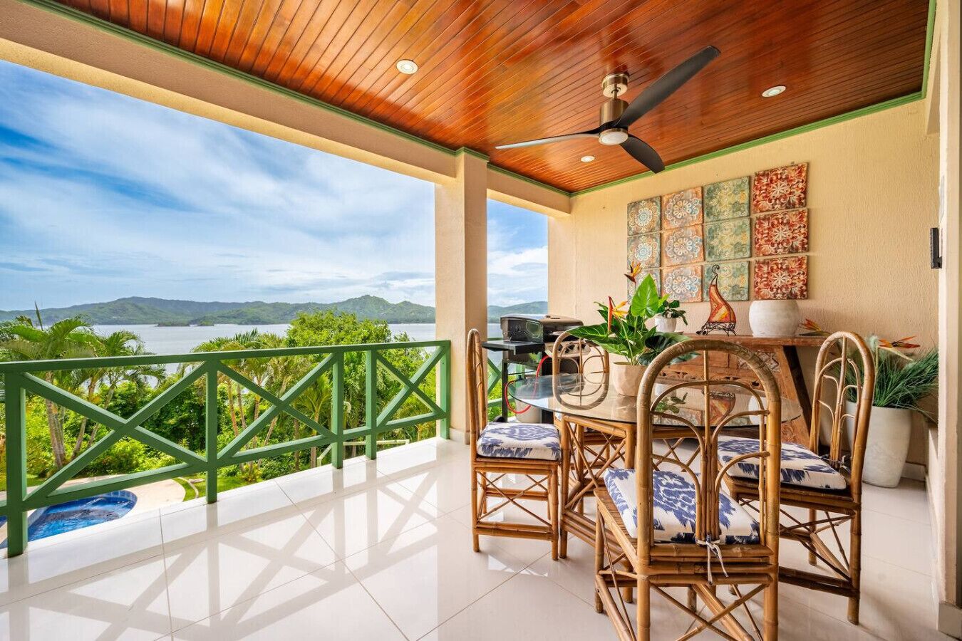 A balcony with a view of a lake and mountains, a table with chairs, and a ceiling fan.