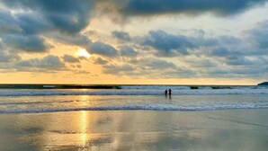 Een strand met twee personen die langs de kust lopen, golven die op het zand breken, en een zonsondergang op de achtergrond.