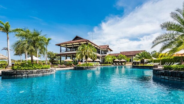 Een resort met een groot zwembad, palmbomen en een twee verdiepingen tellend gebouw met een betegeld dak.