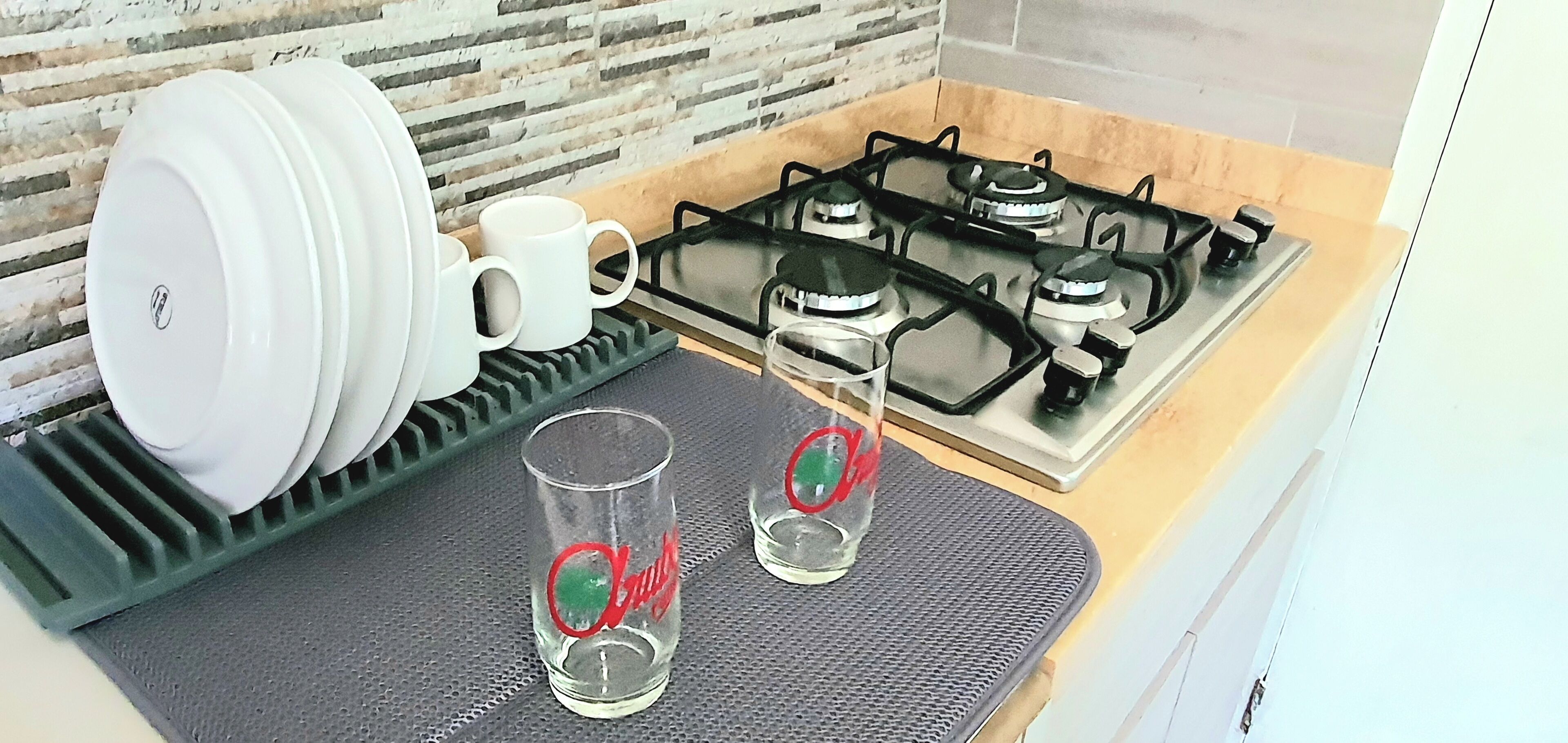 Una cocina con estufa a gas, un escurreplatos con platos y tazas, y dos vasos sobre un tapete.