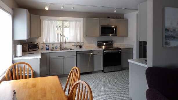 A modern kitchen with a dining area, featuring stainless steel appliances, a window with curtains, and a patterned floor. - Moses Lake Mid-Century Gemstone: Cozy 2-bedroom house in the heart of town. (Moses Lake)