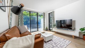 Una sala moderna con un sofá de cuero, una mesa de centro, un televisor de pantalla plana sobre un mueble blanco y un amplio ventanal corredero que da a un balcón con vegetación.