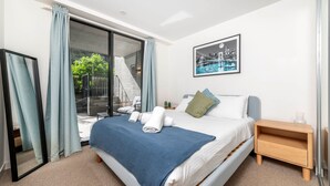 Una habitación de hotel moderna con una cama grande, una mesita de noche, un cuadro con una vista urbana enmarcada y un ventanal corredero que da a un balcón.