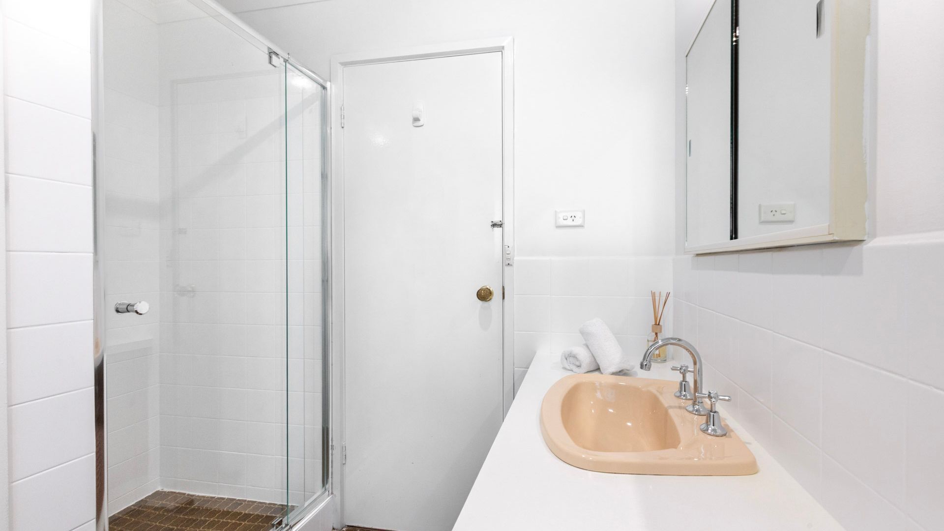 A bathroom with a glass shower enclosure, a beige sink with a mirror above it, and white tiled walls.
