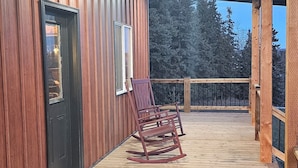 Eine Holzterrasse mit einer schwarzen Tür und Fenstern, einem Schaukelstuhl und einem hölzernen Geländer.