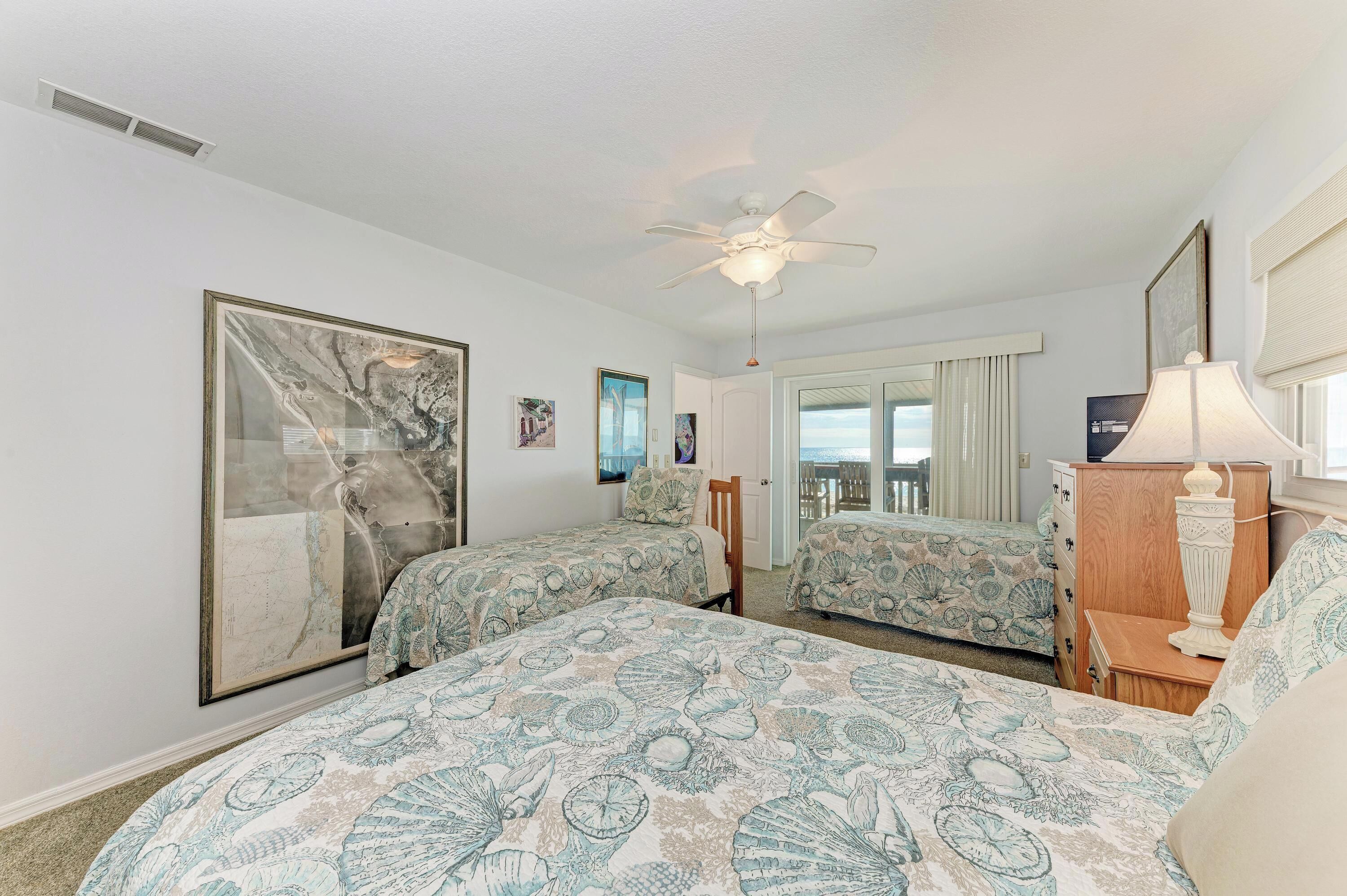 A hotel room with two beds, a ceiling fan, a large framed picture, a lamp, and a view of the ocean through a door.