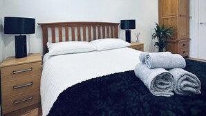 A neatly made bed with white bedding and a dark headboard, a wooden nightstand with a lamp, a wooden wardrobe, and a rolled-up towel on the floor.