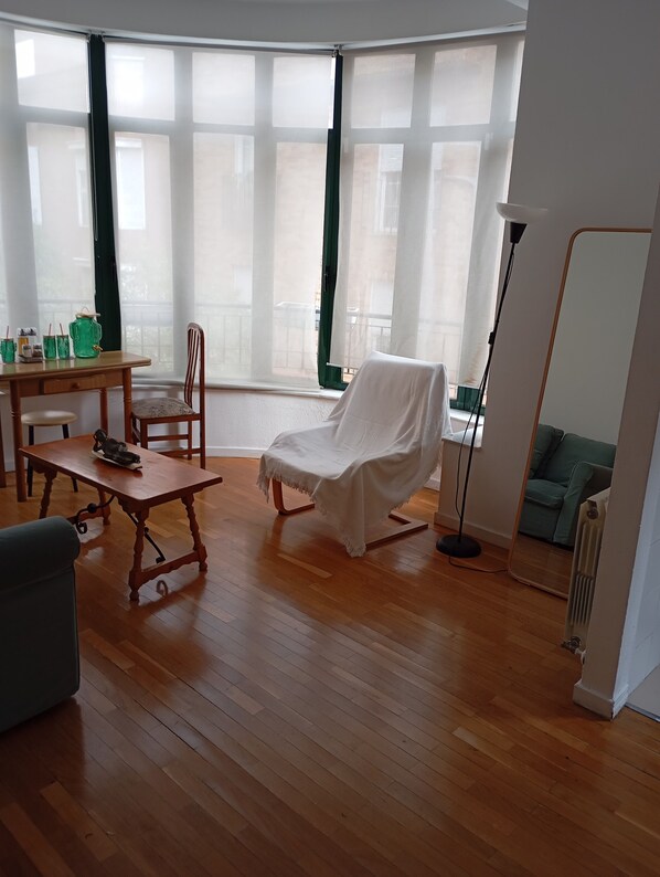 Une pièce avec un plancher en bois, une chaise blanche, une table avec une chaise, un canapé vert et une grande fenêtre avec des rideaux.