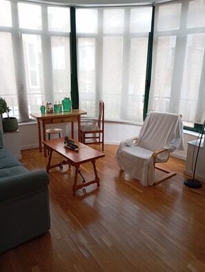 Un salon avec un plancher en bois, un canapé vert, une table à manger avec des chaises et un fauteuil blanc recouvert d’un tissu.