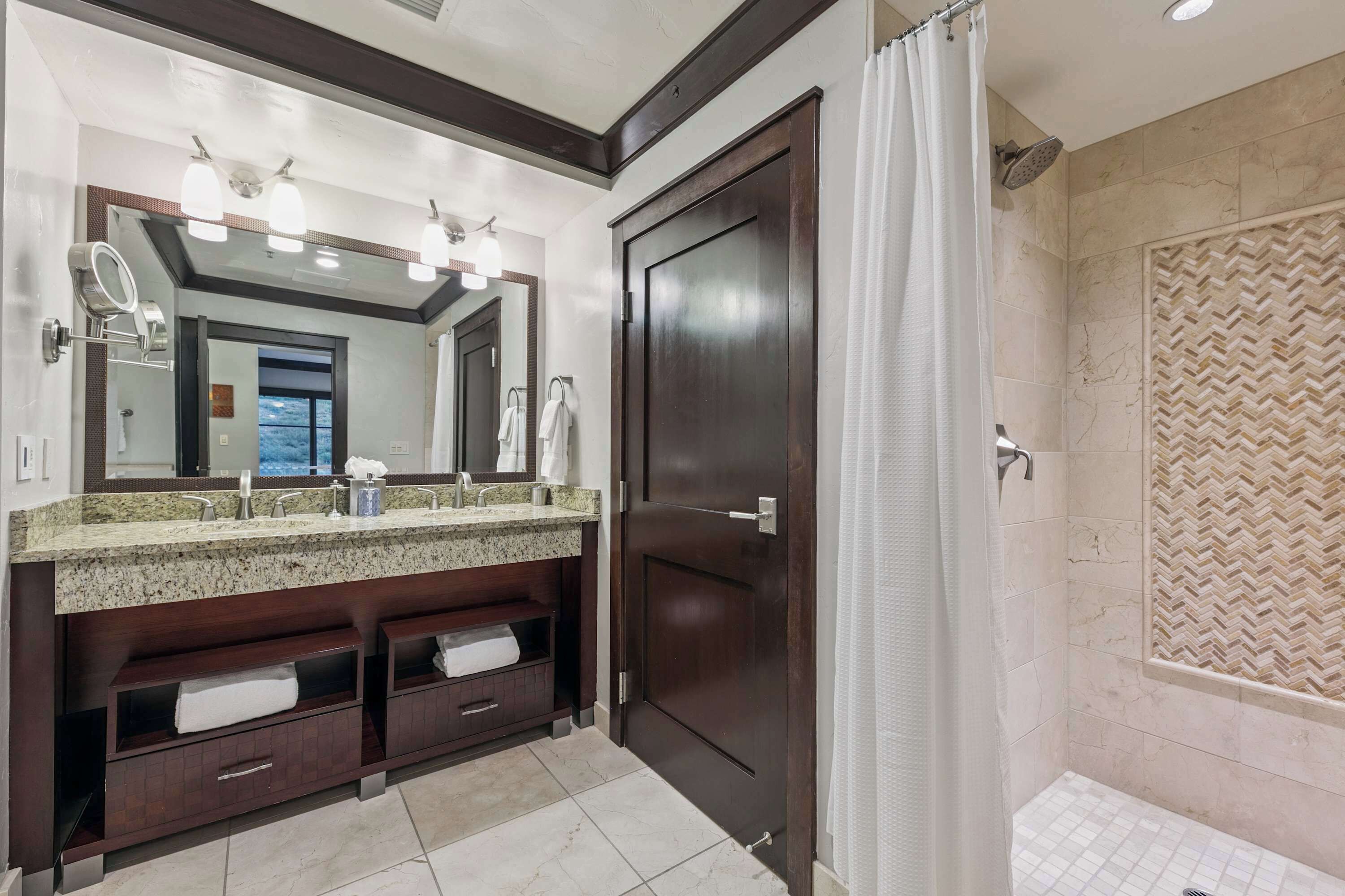 A bathroom with a dark wooden vanity, a large mirror, and a shower with a herringbone tile pattern.