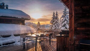 Une terrasse en bois avec des tables et des chaises, des arbres couverts de neige et une chaîne de montagnes en arrière-plan.