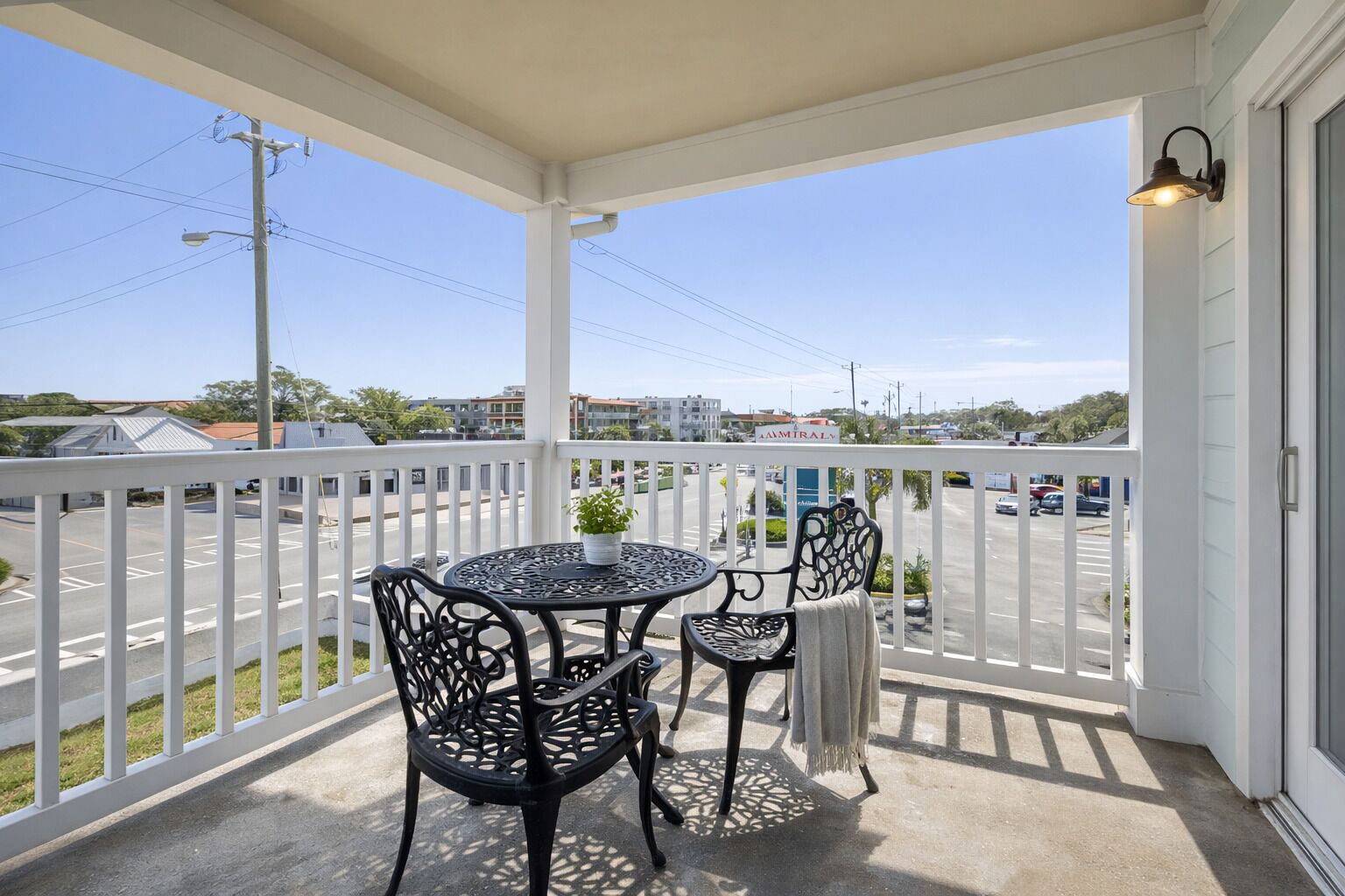 A covered balcony with a table and chairs, overlooking a street with buildings and cars.