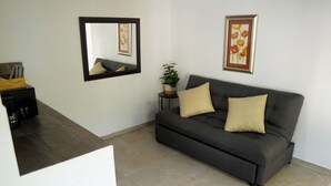 A living room with a dark grey sofa, two beige cushions, a small side table with a potted plant, a framed picture, and a mirror on the wall. - Campestre La Fuente  (Monterrey)