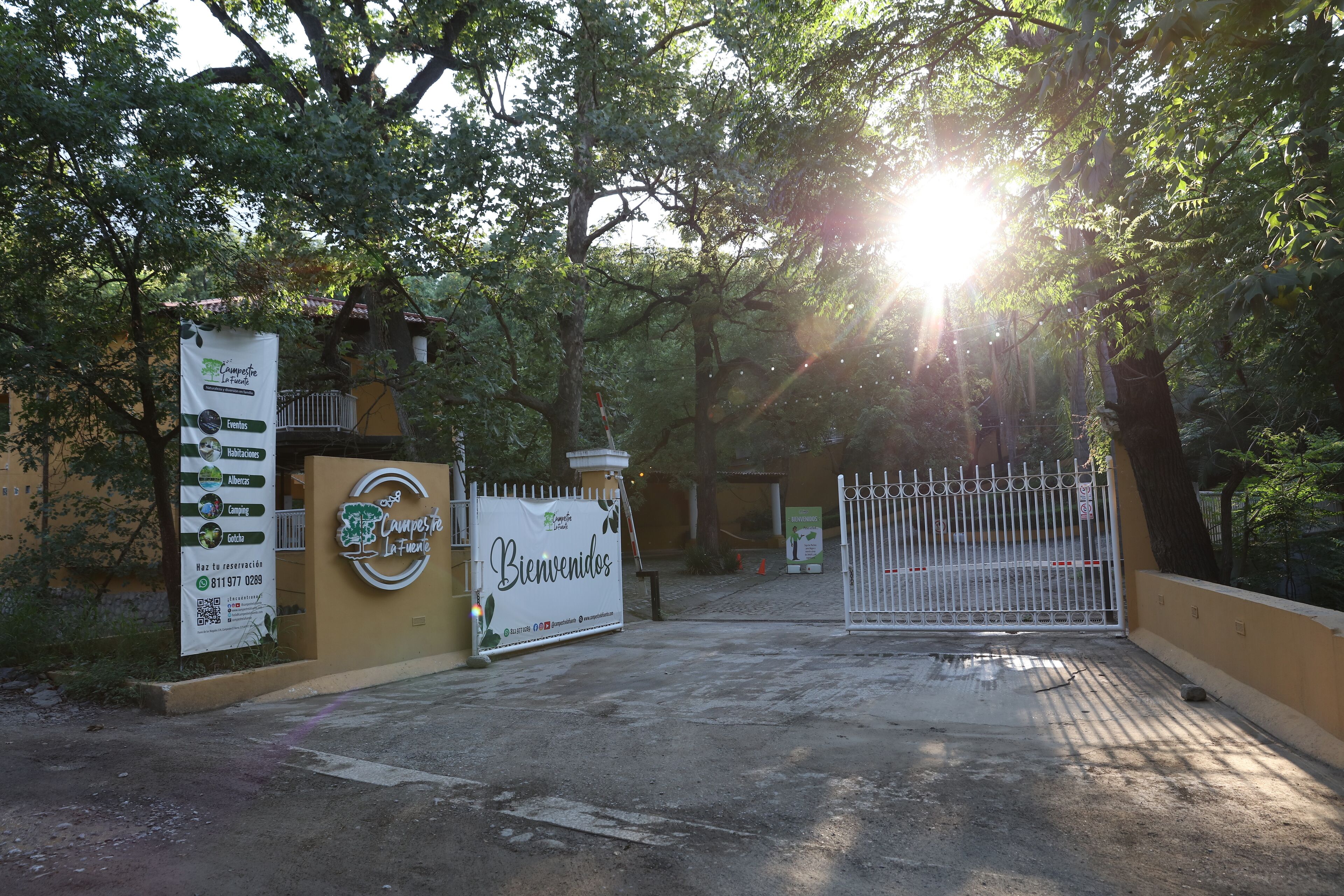 Una entrada protegida por una reja con un letrero de bienvenida, rodeada de árboles y un ambiente soleado.