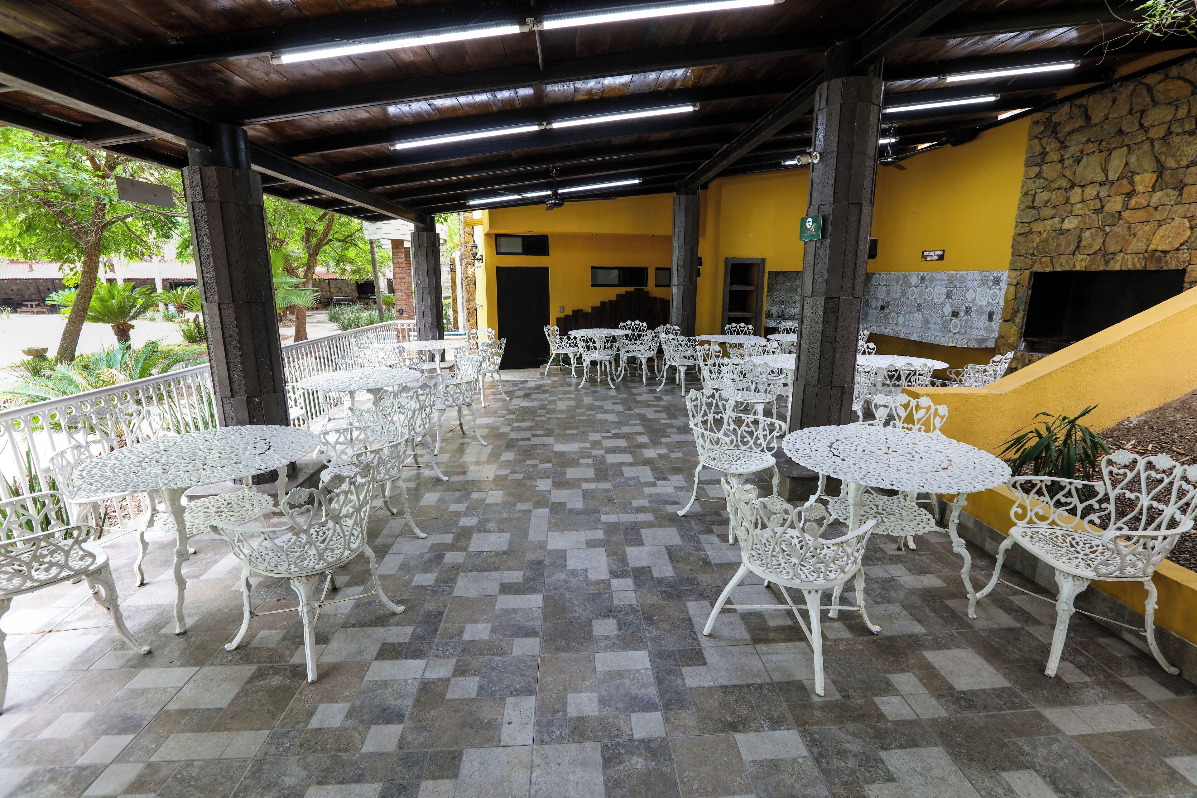 Un patio al aire libre con mesas y sillas de metal blanco, un muro de piedra y una pared amarilla con chimenea.