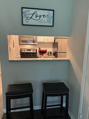 Una cocina compacta con una encimera, un microondas, un tostador y un cuenco de fruta. Hay dos taburetes debajo de la encimera.