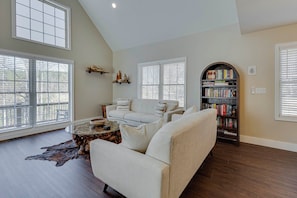 Un salon spacieux avec un plafond haut, de grandes fenêtres équipées de stores, un canapé blanc, une table basse et une bibliothèque.