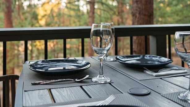 Una mesa de comedor al aire libre con platos, cubiertos y una copa de vino, rodeada de árboles altos.