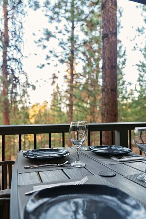 Una mesa de comedor al aire libre con platos, cubiertos y una copa de vino, rodeada de árboles altos.