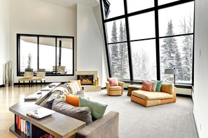 A spacious living room with a fireplace, large windows, and a view of snow-covered trees. - KBM Resorts| Walk to Slopes, Sports Court, Private Home Theater, Sleeps 17, Outdoor Hot Tub PCO-184M (Park City)