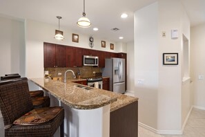 A kitchen with granite countertops, stainless steel appliances, dark wood cabinets, and a central island with seating. - Fairway Villas Waikoloa | Golf Course View 2 BR | Free Car w/ 6+ Nts | FVW-L32 (Waikoloa Village)