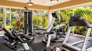 Un gimnasio con diferentes equipos de ejercicio, ventanas amplias y vistas al exterior con vegetación.