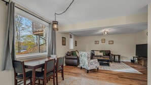 A living room with a dining area, wooden flooring, a large window with a view of trees, a sofa set, and a television. - Lake House—Unit B (East Lyme)