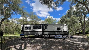 A recreational vehicle (RV) parked in a wooded area with picnic tables and a grill. - Cozy 1-bedroom mobile home in beautiful Spring Hill with AC (Spring Hill)