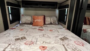 A bed with a Paris-themed duvet cover, a headboard with built-in shelves, and a window with blinds. - Cozy 1-bedroom mobile home in beautiful Spring Hill with AC (Spring Hill)