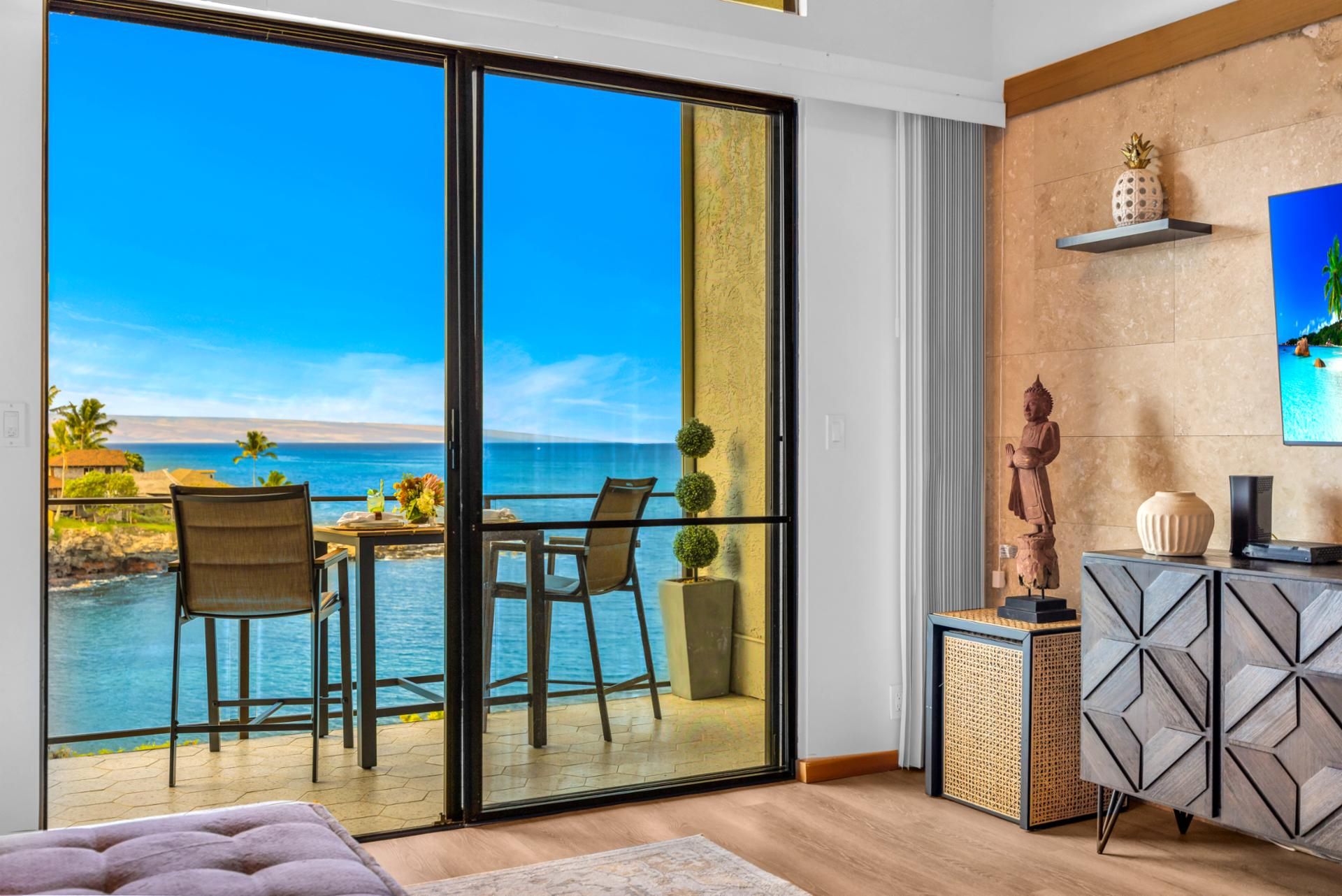 A room with a large sliding glass door leading to a balcony with a view of the ocean and a tropical landscape.