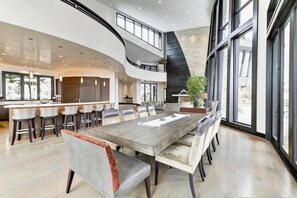 A spacious dining area with a long table, multiple chairs, and a high ceiling with large windows.
