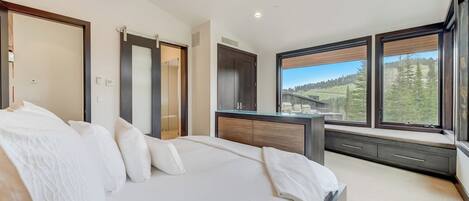 A bedroom with a large bed, a dresser, a vanity, and a window with a view of trees and buildings.