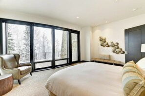 A bedroom with a large bed, a chair, a bench, and a view of trees through large windows.