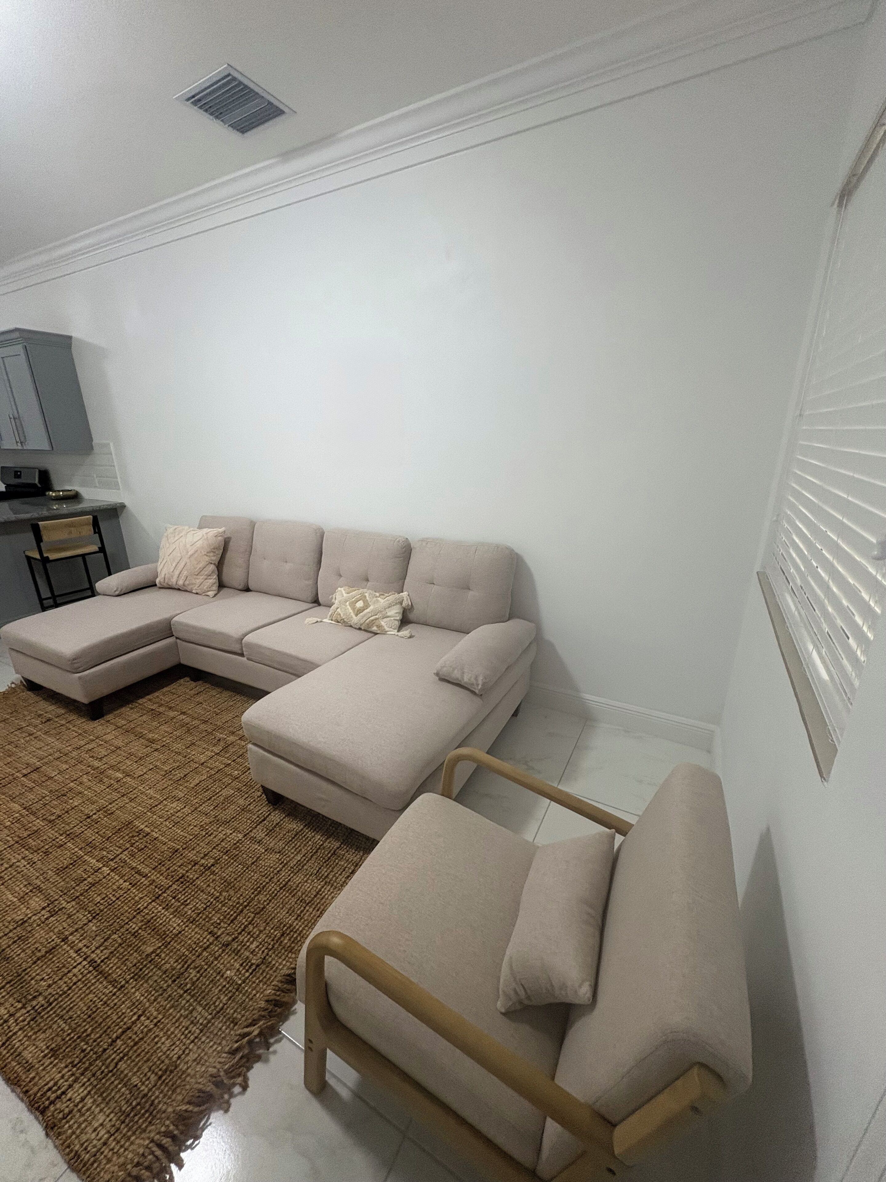 Una sala moderna con un sofá seccional color beige, una pequeña silla de madera y un tapete tejido marrón.